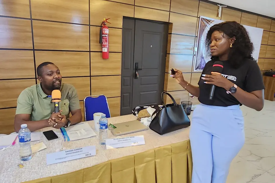 A facilitator and a seated participant each hold a microphone during a session at a conference table.