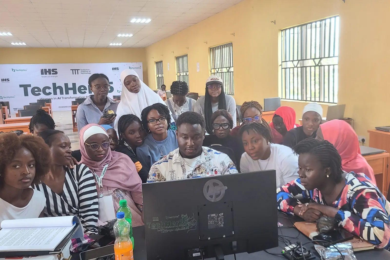 Students gather around their instructor and a shared monitor in a classroom.