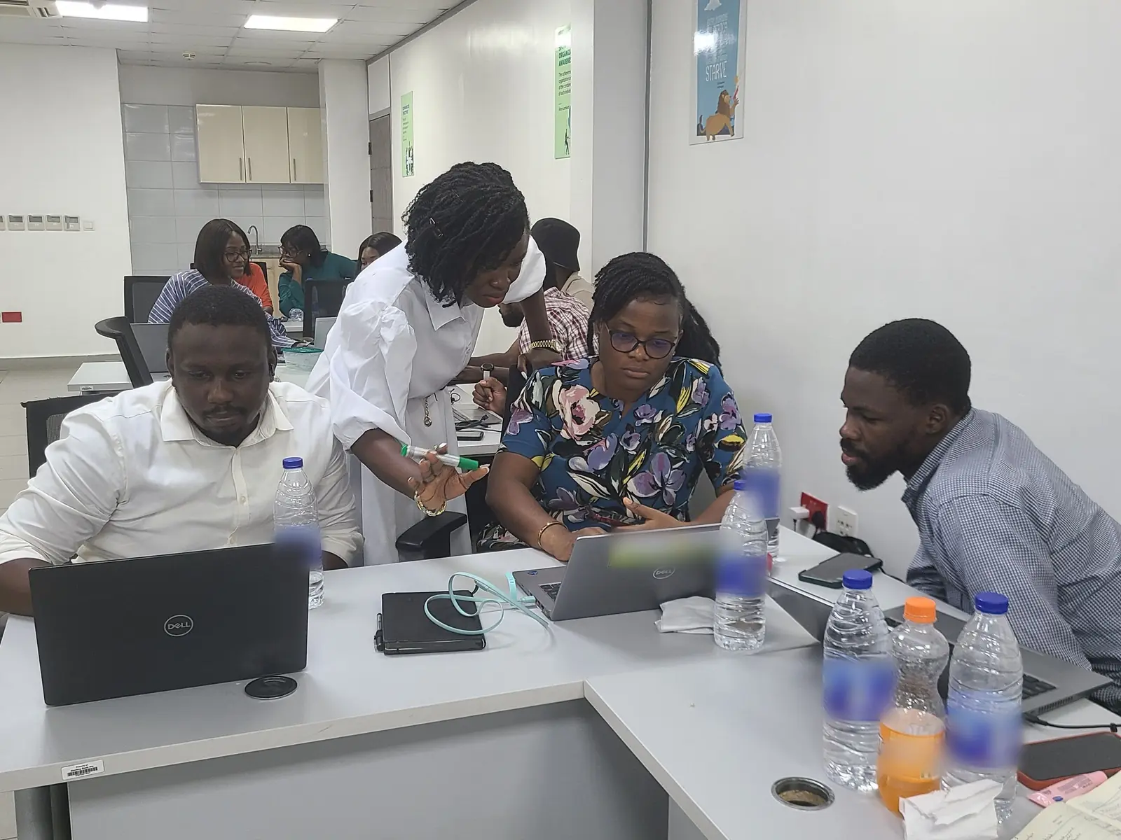 A facilitator leans over a shared table to help a participant with a laptop while colleagues work beside them.