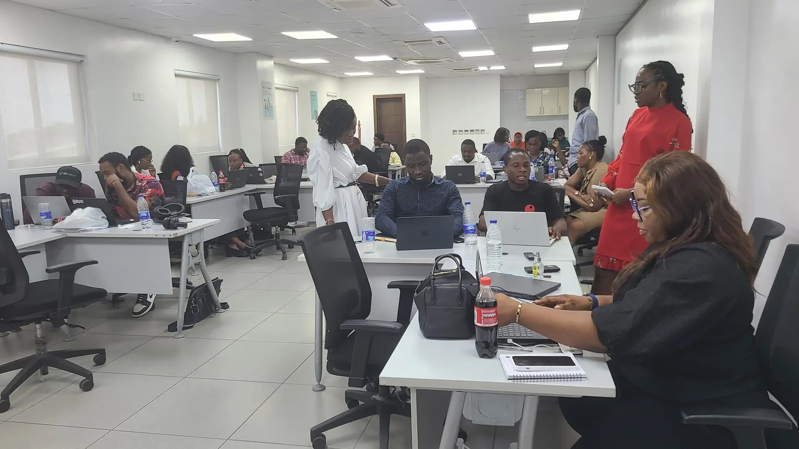Participants work at laptops across a large open training room while a facilitator speaks with a learner.